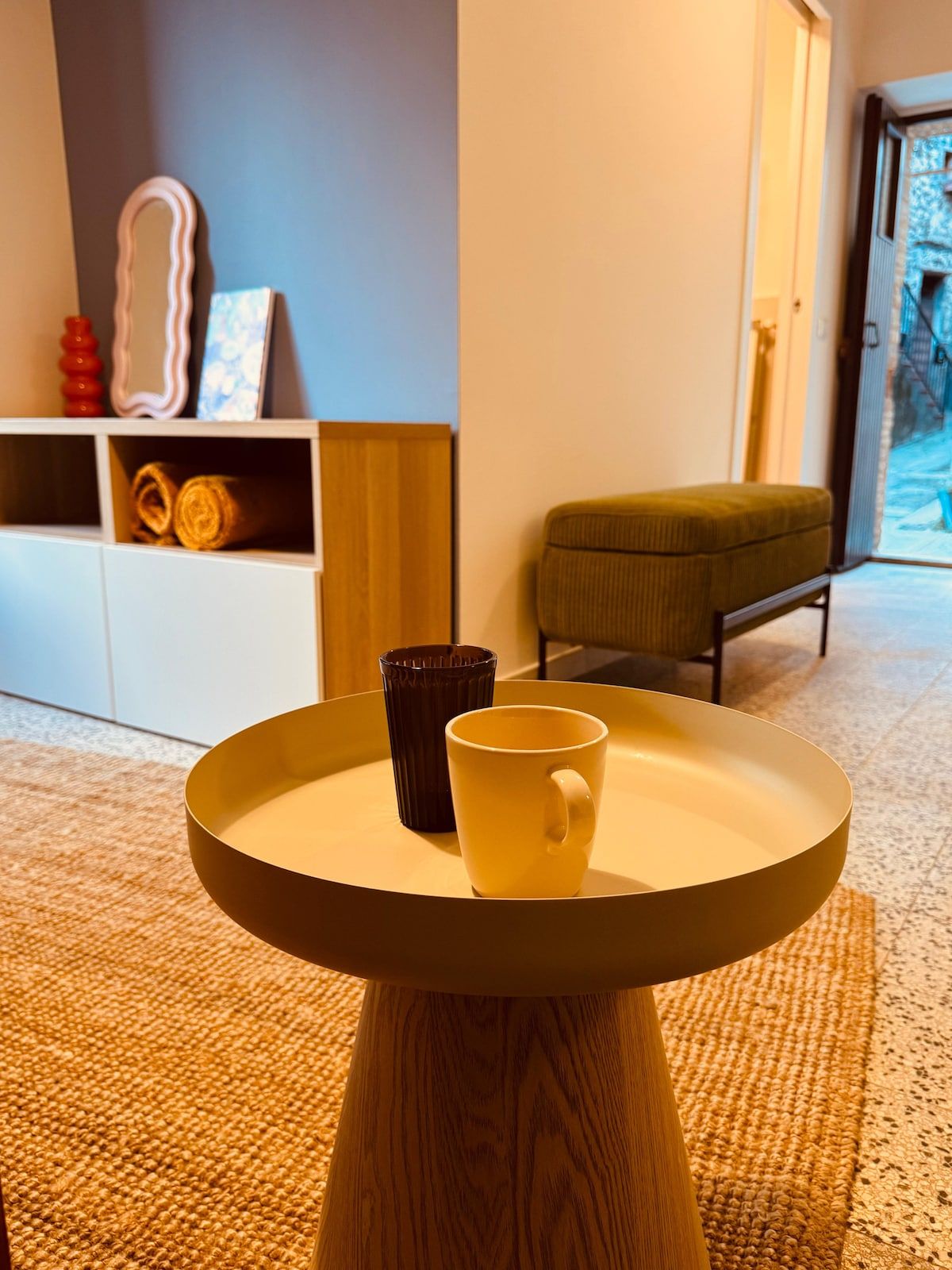 Un tavolo rotondo in legno con un vassoio che contiene due tazze, un armadietto in legno con oggetti decorativi e una panca verde in una stanza con pavimento tappezzato.