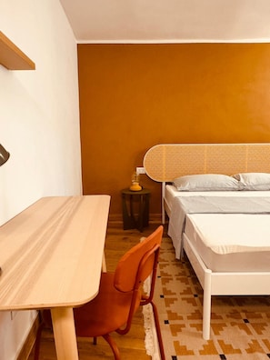 Ein kleines Schlafzimmer mit einem Holztisch, einem einzelnen orangefarbenen Sessel, einem Bett mit weißem Kopfteil und einem kleinen runden Tisch mit einer Lampe.