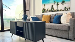 Ein modernes Wohnzimmer mit einem grauen Sofa, einer schwarzen und grauen Unterhaltungsecke und einem großen Fenster mit Blick auf den Himmel und einem Gemälde mit einer Strandansicht.