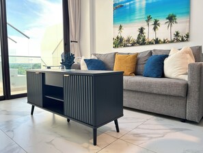 A modern living room with a grey sofa, a black and grey entertainment unit, and a large window with a view of the sky and a painting of a beach scene. - Cana Rock Star/Golf and Pool/Hard Rock Area (Punta Cana)
