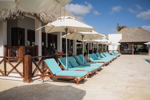 A row of wooden lounge chairs with white umbrellas, a thatched-roof building, and a pool area. - Cana Rock Star/Golf and Pool/Hard Rock Area (Punta Cana)