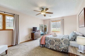Ein Schlafzimmer mit Bett, Fernseher, Schminktisch und Deckenventilator, ausgestattet mit großen Fenstern.
