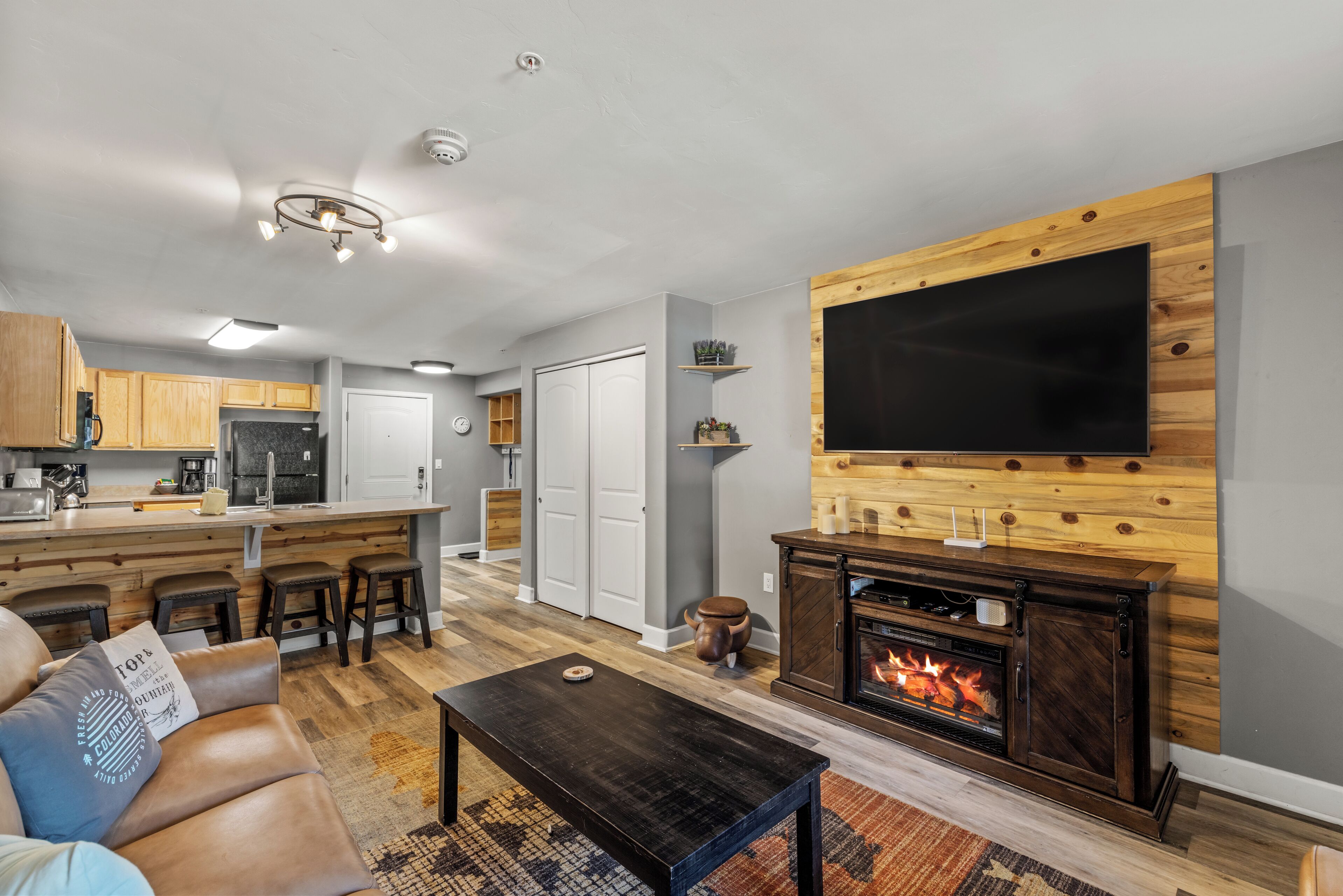 Un salon moderne doté d’une cheminée, d’un grand téléviseur à écran plat fixé à un mur en bois, d’un coin cuisine avec des meubles en bois et d’un coin repas avec des tabourets de bar.