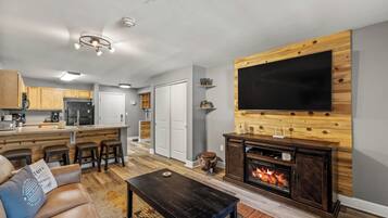 Un salon moderne doté d’une cheminée, d’un grand téléviseur à écran plat fixé à un mur en bois, d’un coin cuisine avec des meubles en bois et d’un coin repas avec des tabourets de bar.