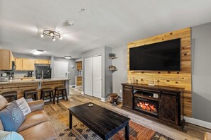 Ein modernes Wohnzimmer mit Kamin, einem großen Flachbildfernseher an einer Holzwand, einer Küchenzeile mit Holzschränken und einem Essbereich mit Barhocker.