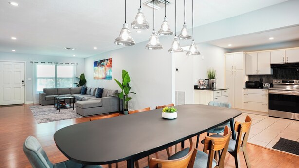 Uma cozinha moderna com eletrodomésticos de aço inoxidável, uma sala de jantar com mesa escura e cadeiras azuis, e uma sala de estar com sofá cinza e uma janela grande.
