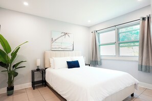 Een slaapkamer met een bed, een nachtkastje, een plant in pot, een raam met gordijnen en een aan de muur hangend schilderij.