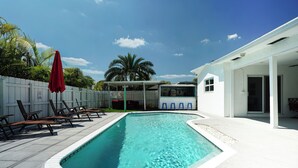 Piscina com espreguiçadeiras e guarda-sóis, cerca branca e uma casa ao fundo.