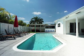 A swimming pool with lounge chairs and umbrellas, a white fence, and a house in the background.