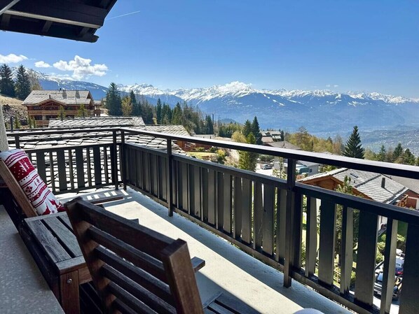 A balcony with a wooden bench and a railing, overlooking a mountain landscape with snow-capped peaks. - Sunny Alpine Apartment - 180 View (Lens)