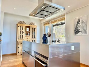 A modern kitchen with a stainless steel island, wooden cabinets, and a large window. - Sunny Alpine Apartment - 180 View (Lens)