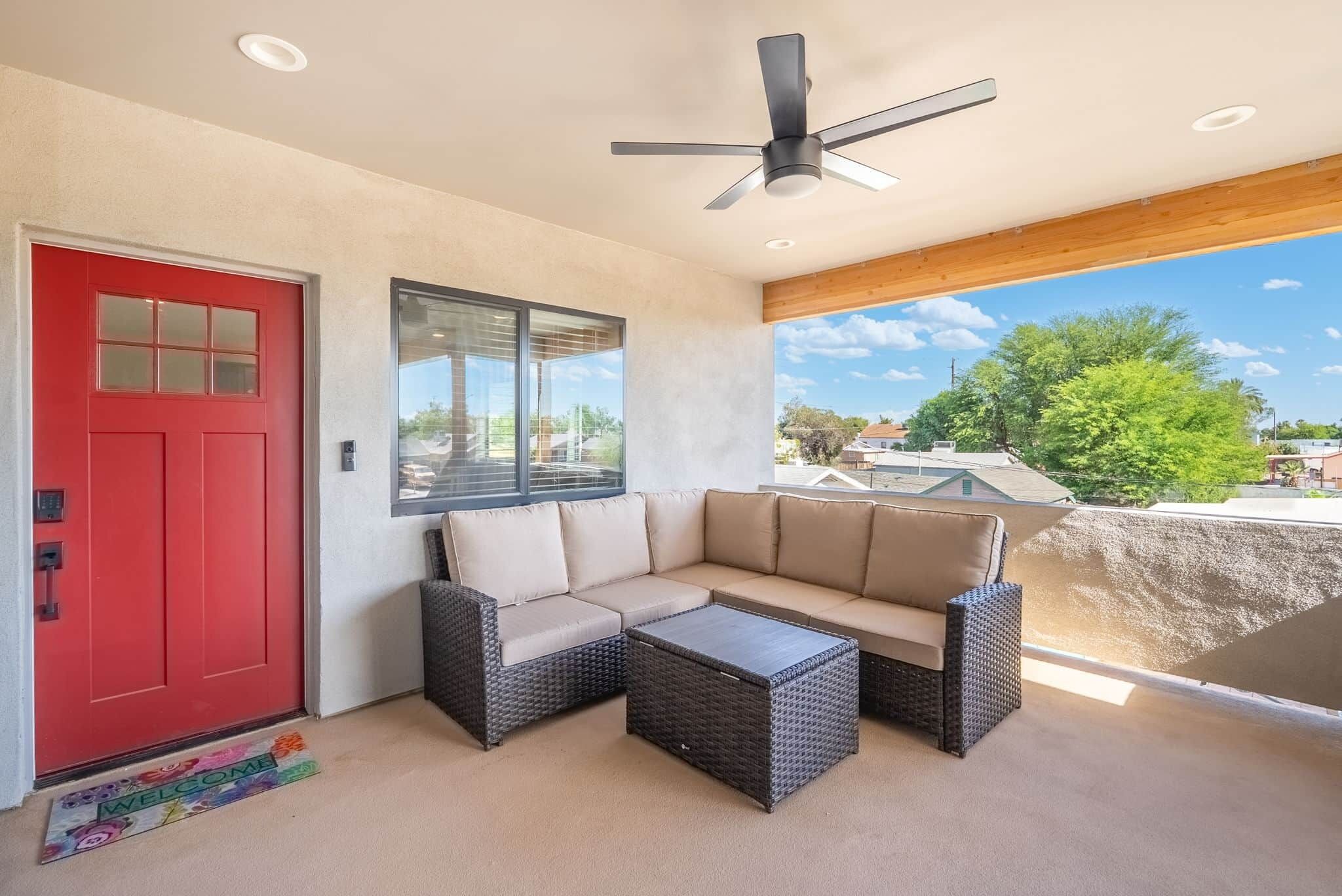 A covered patio with a red door, a beige sofa, a small table, and a ceiling fan.