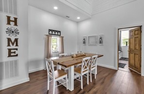 A dining area with a wooden table and white chairs, a window with curtains, and a door leading to another room.