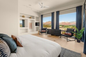 Ein modernes Hotelzimmer mit einem großen Bett, einem Schreibtisch, zwei Stühlen, einem Fernseher und Blick auf eine gebirgige Landschaft.