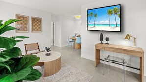 Uma sala de estar moderna com uma mesa de centro redonda, uma TV fixada na parede, uma escrivaninha de madeira e um espaço de jantar com cadeiras.