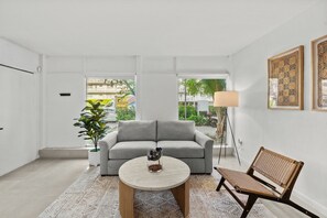 Ein modernes Wohnzimmer mit einem grauen Sofa, einem runden Couchtisch, einem Korbstuhl und einer Topfpflanze.
