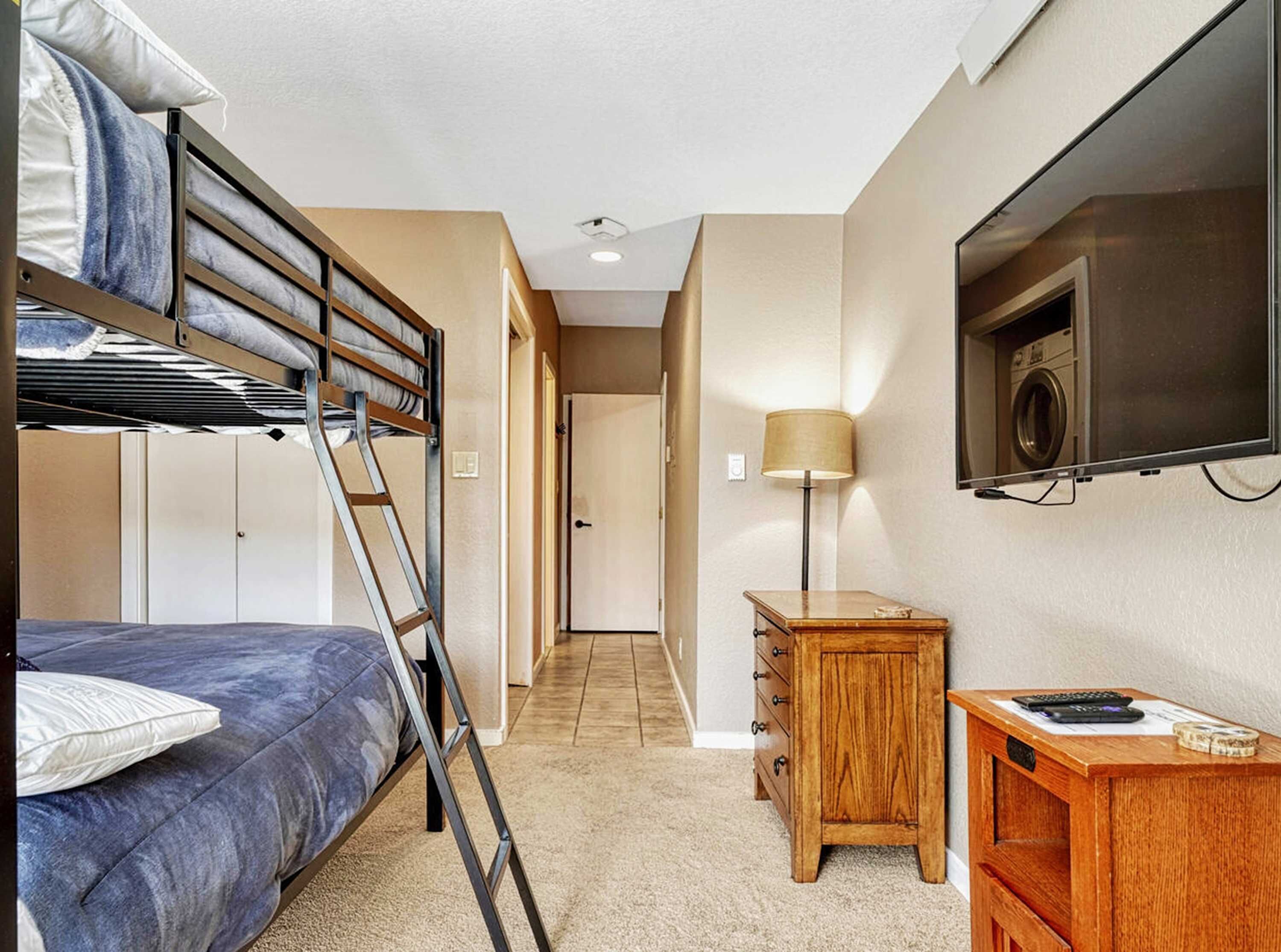 A compact hotel room with bunk beds, a small wooden dresser, a desk with a lamp, and a TV mounted on the wall.