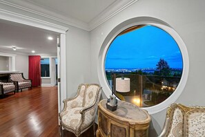 Ein Zimmer mit einem großen Rundfenster, zwei verzierten Stühlen, einem kleinen Tisch und Blick auf eine Stadtlandschaft im Abenddämmerung.
