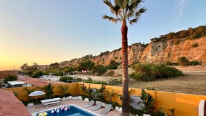A swimming pool with lounge chairs and a palm tree, surrounded by a herringbone-patterned patio and a view of a cliff. - CASA RIBA - between ocean & countryside with pool (caparica)