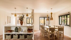 A dining area with a long table and chairs, a white shelving unit with baskets, and hanging pendant lights. - CASA RIBA - between ocean & countryside with pool (caparica)