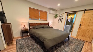 A bedroom with a bed, a nightstand, a lamp, a guitar, a window with blinds, a wall-mounted air conditioner, a door, and a small plant. - Cozy 2-bedroom house in tranquil Prescott Valley with AC (Prescott Valley)