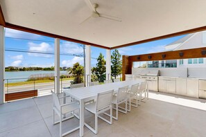 A modern outdoor dining area with a white table and chairs, a ceiling fan, and a view of a body of water and trees. - 7 Bedroom Waterfront Rehoboth beach house with private pool (Rehoboth Beach)