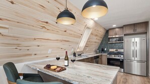 A kitchen with wooden walls, a marble countertop, a dining area with chairs, and stainless steel appliances. - Brand New A-Frame! Panoramic Mt Views, Hot Tub, Speakeasy, EV Charger (Florissant)