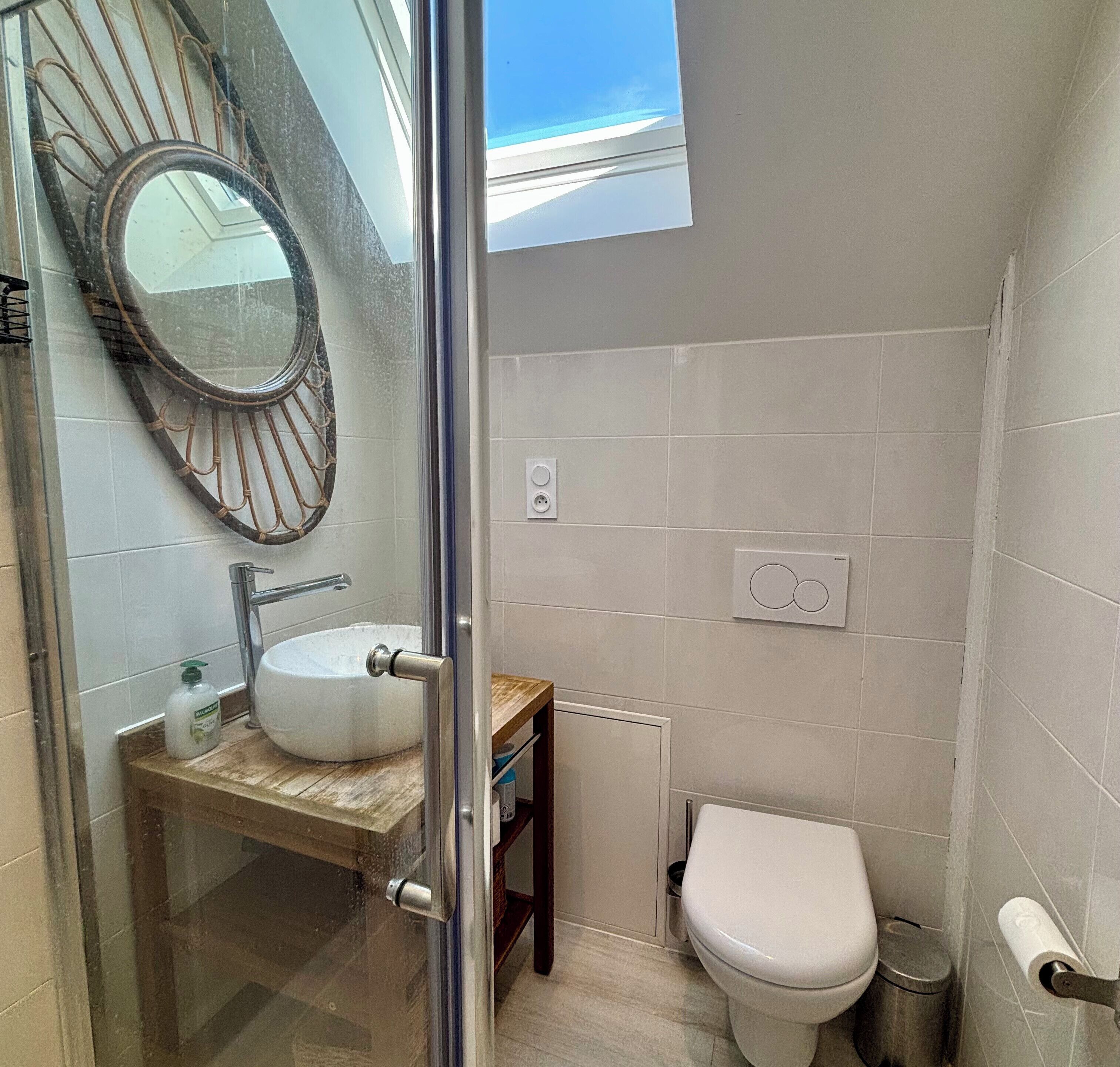 A bathroom with a glass shower enclosure, a round mirror, a white sink, and a toilet.