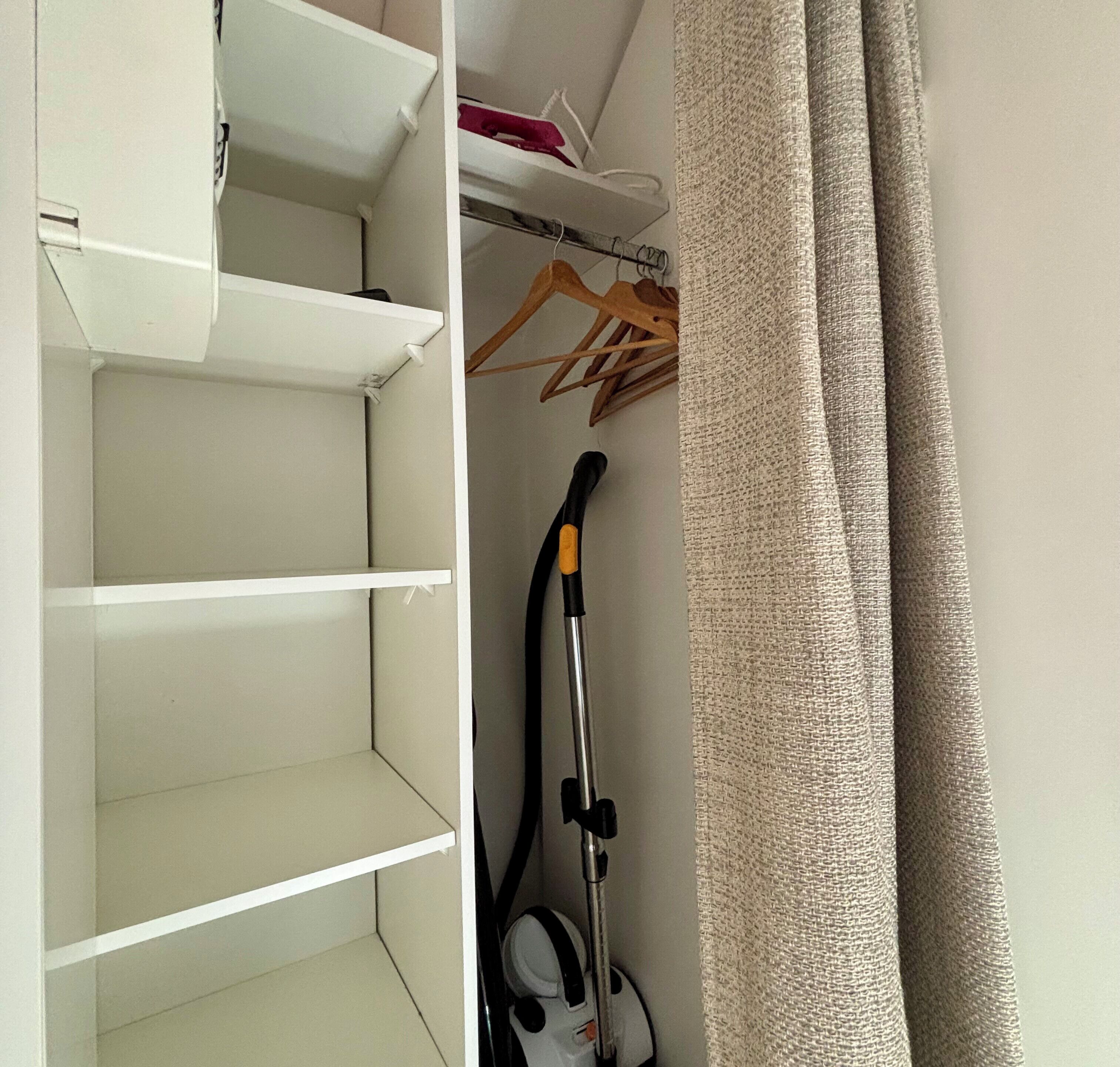 Um armário com prateleiras abertas, um aspirador de pó e uma cortina.