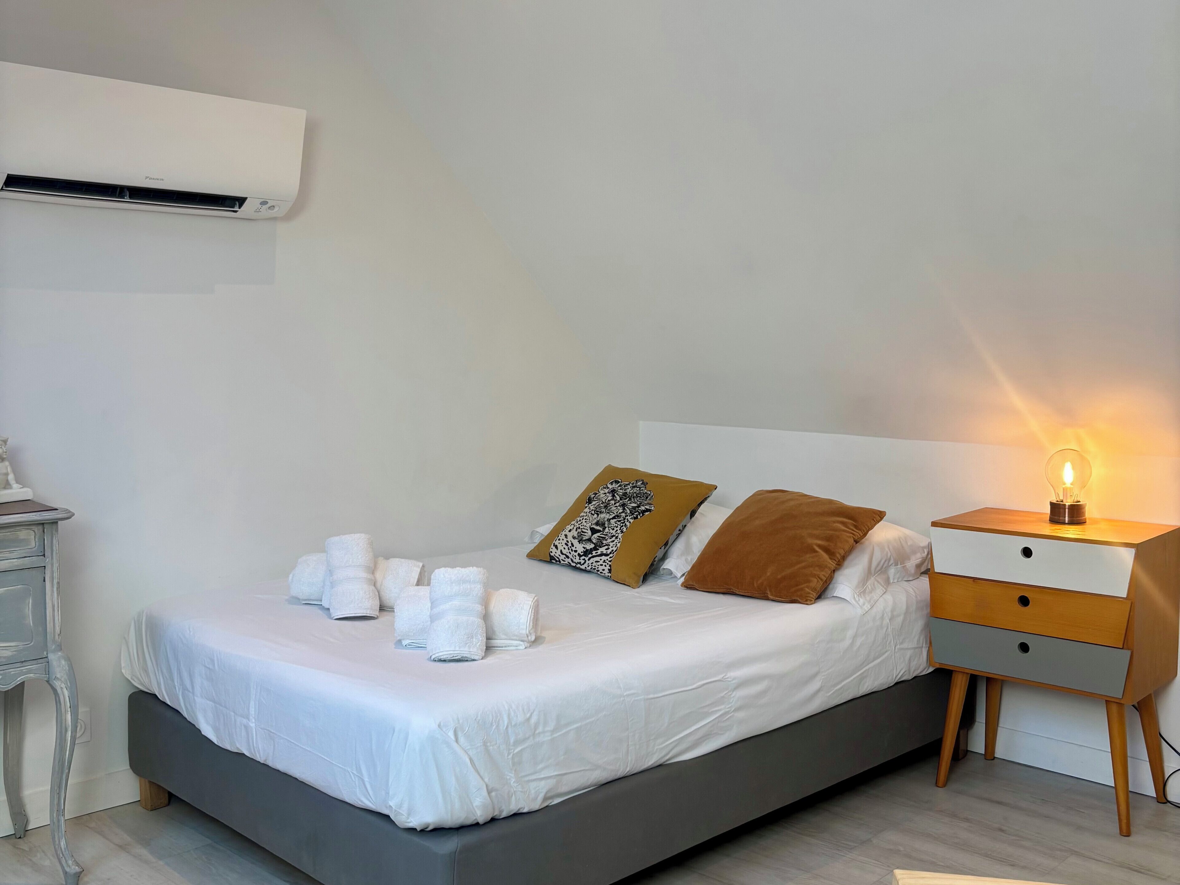 A neatly made bed with white linens, a bedside table with a lamp, and a wall-mounted air conditioner.