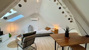 Ein gemütliches Schlafzimmer im Loft-Stil mit Dachfenster, einem Bett, einem Nachttisch mit Lampe, einem Sessel und einem Esstisch.