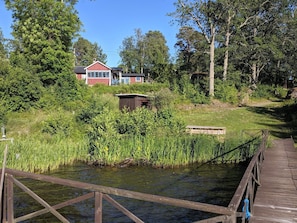 A wooden dock extending over a calm body of water, surrounded by lush greenery and a small wooden structure.