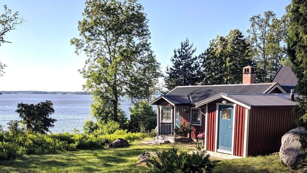 A cabin by a lake with a clear view of the water, surrounded by trees and greenery. - 4 Star Holiday Home in Eskilstuna (ESKILSTUNA)