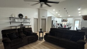 Ein modernes Wohnzimmer mit zwei dunklen Sofas, einem Deckenventilator und einer Küche im Hintergrund.