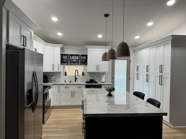 Una cocina moderna con armarios blancos, un refrigerador de acero inoxidable, una isla de mármol, luces colgantes y salpicadero con baldosas hexagonales.