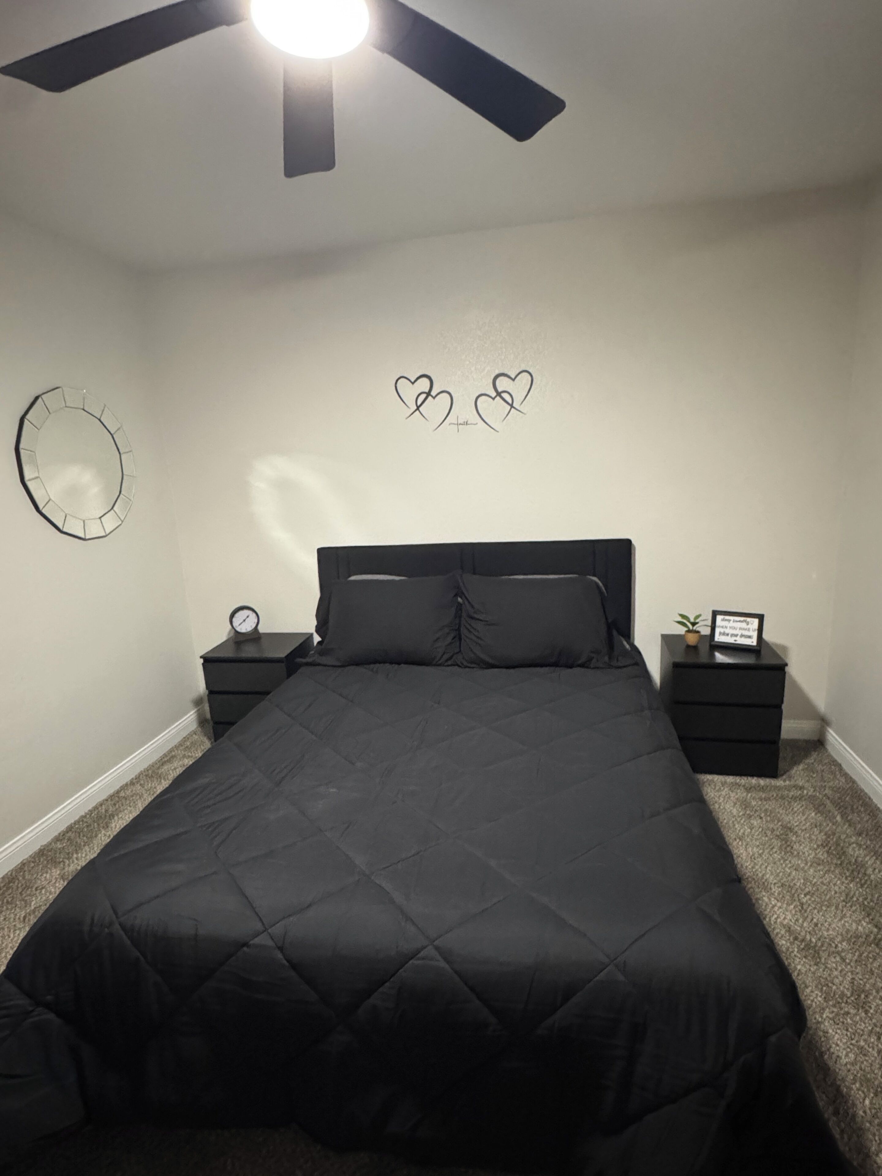 Een slaapkamer met een bed, twee nachtkastjes, een wandklok, een lijst met een citaat, een plafondventilator en een wanddecoratie.