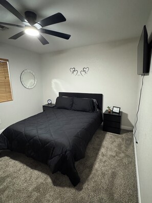 A bedroom with a bed, a nightstand, a wall clock, a ceiling fan, a TV, a mirror, and wall art.