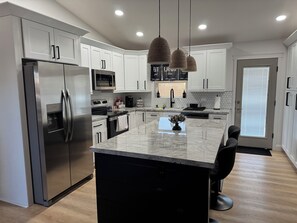 A modern kitchen with stainless steel appliances, a marble island, pendant lighting, and a view to the outside.