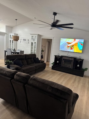 A modern living room with a sectional sofa, a flat-screen TV mounted on the wall, a ceiling fan, and a fireplace.