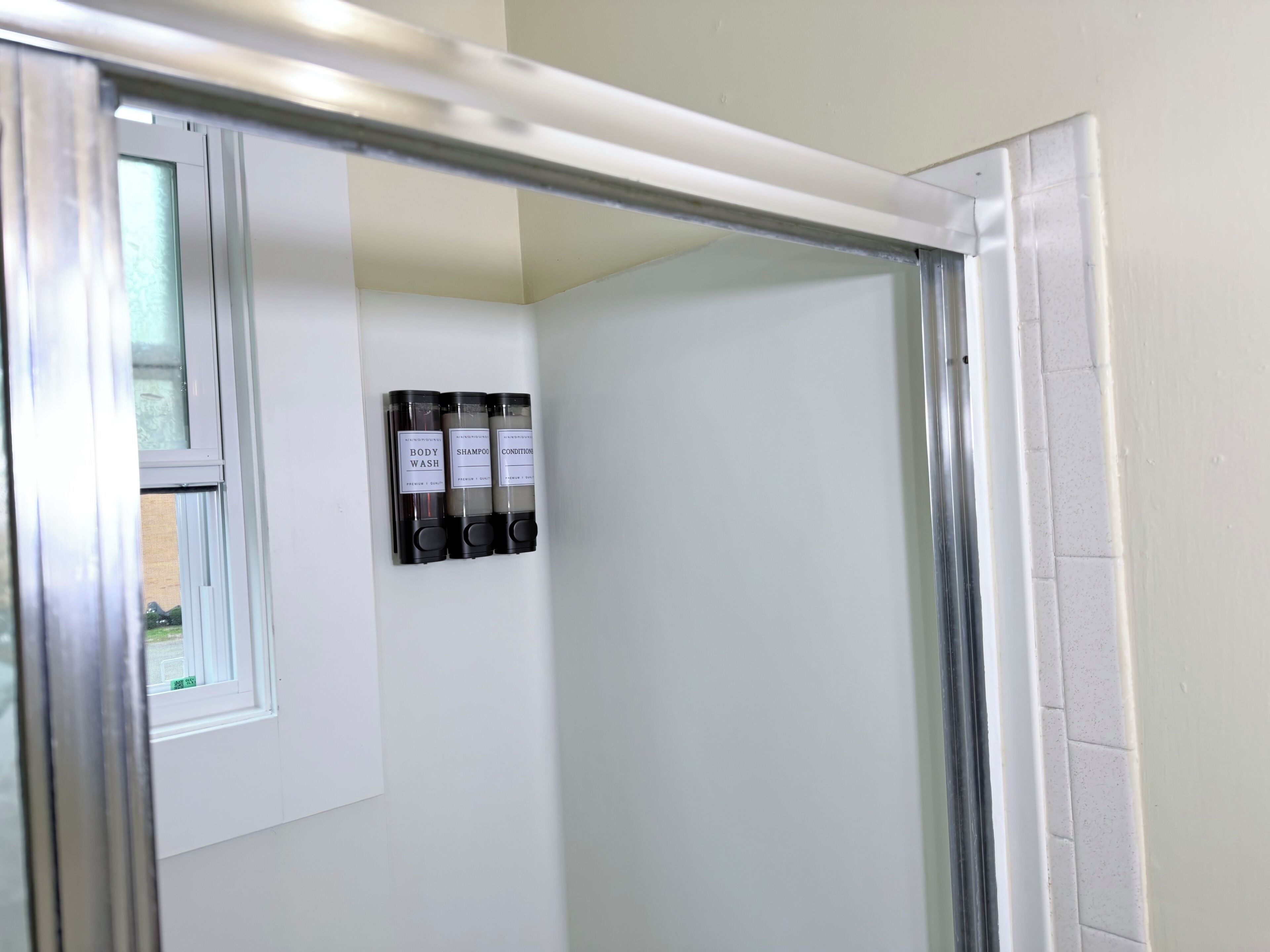 A bathroom with a shower, a window, and three bottles of shampoo and conditioner on the wall.