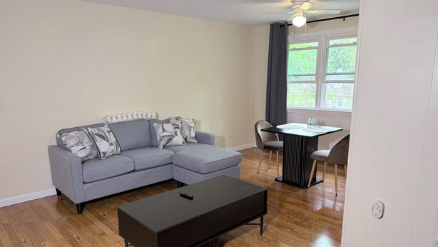 Ein Wohnzimmer mit einem Ecksofa, einem Couchtisch, einem Esstisch mit Stühlen und einem Fenster mit Vorhängen.