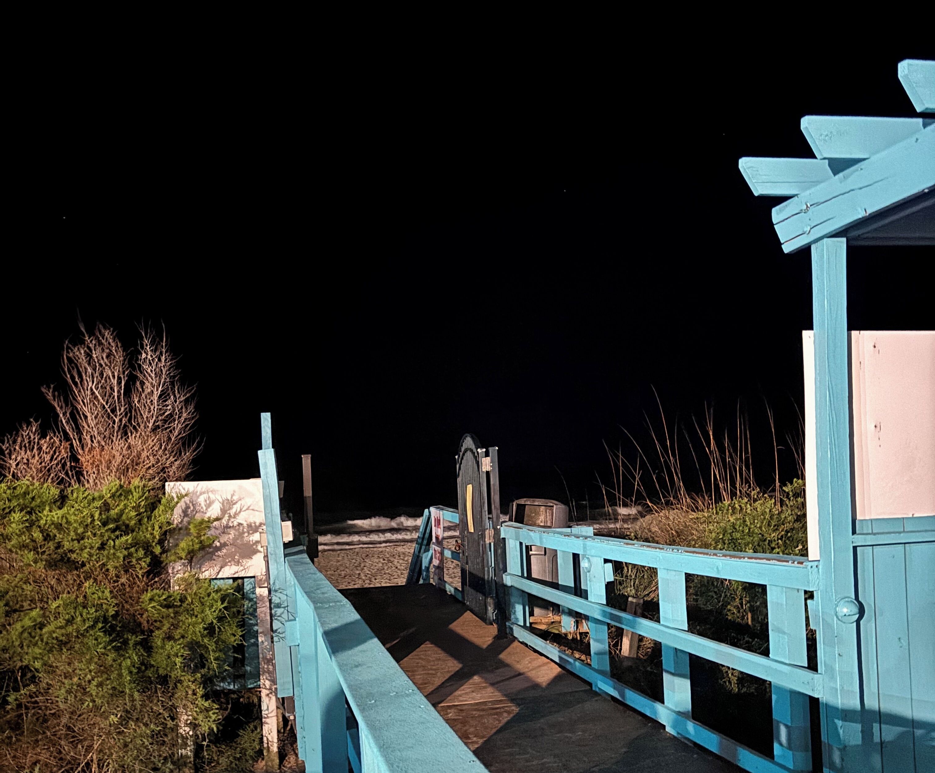 A wooden bridge with railings, leading to a building with a dark roof.