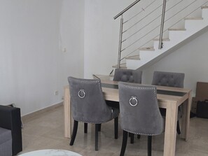 Ein Essbereich mit einem Holztisch und vier grauen, gepolsterten Stühlen mit Metallringen. Im Hintergrund ist eine Treppe mit Metallgeländer zu sehen.