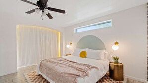 Ein Schlafzimmer mit einem Bett, einem Deckenventilator, Nachttischlampen, einem Fenster und einer kleinen Pflanze auf einem Nachttisch.
