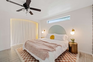 Ein Schlafzimmer mit einem Bett, einem Deckenventilator, Nachttischlampen, einem Fenster und einer kleinen Pflanze auf einem Nachttisch.
