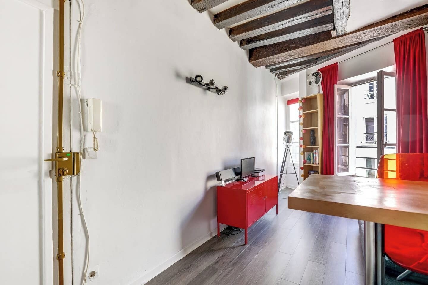 A room with a wooden ceiling, a red desk with a computer, a bookshelf, and a window with red curtains.