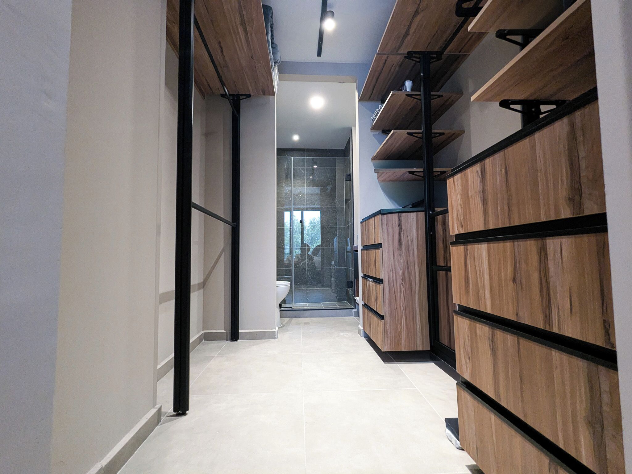 A hallway with wooden shelving units, a glass door leading to a bathroom, and a tiled floor.