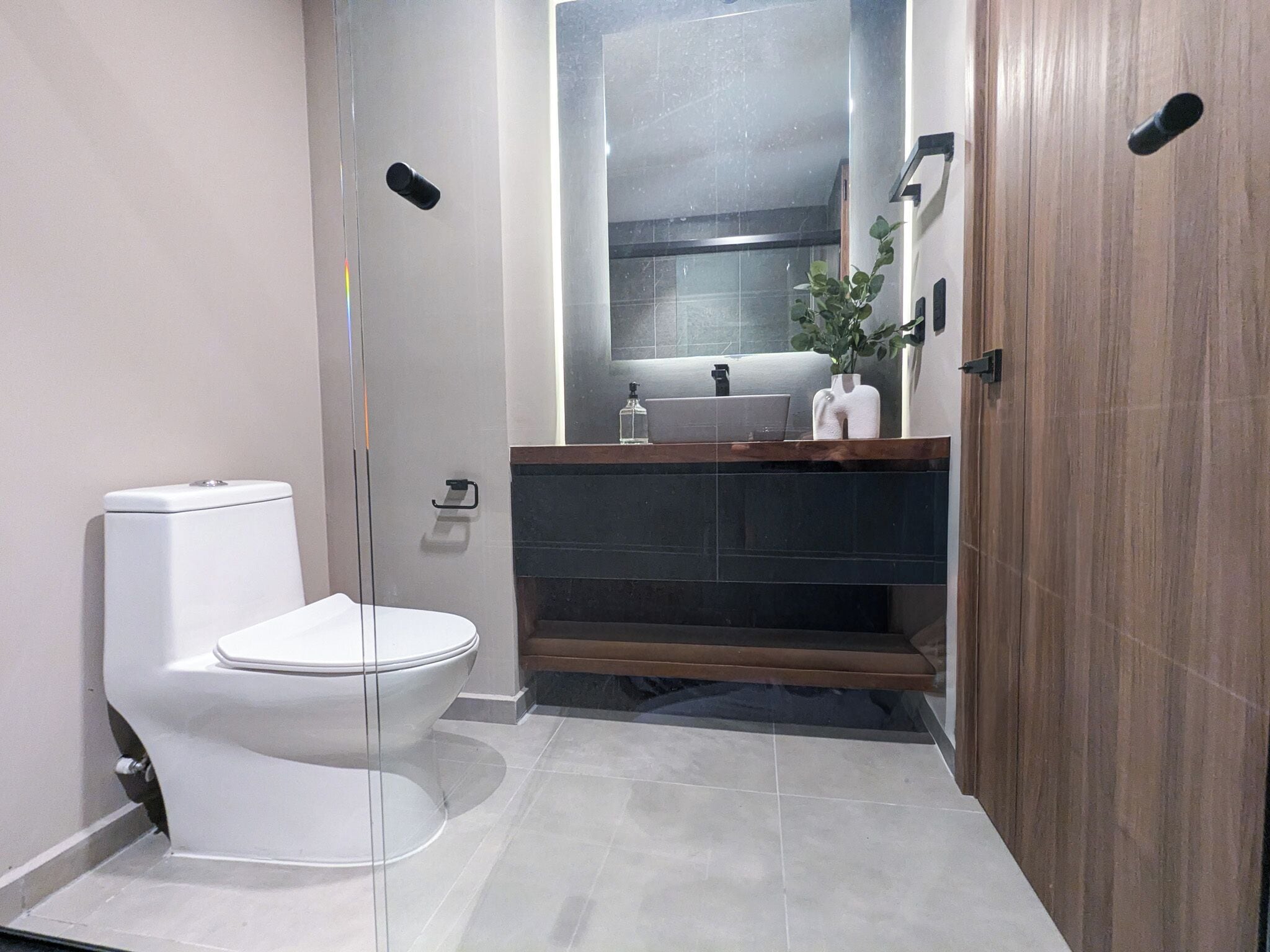 Un baño moderno con inodoro blanco, tocador de madera y espejo sobre el lavamanos.