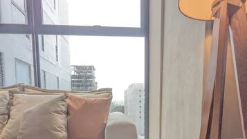 Un salón moderno con sofá, almohadones, una lámpara de pie y vistas a edificios a través de amplias ventanas.