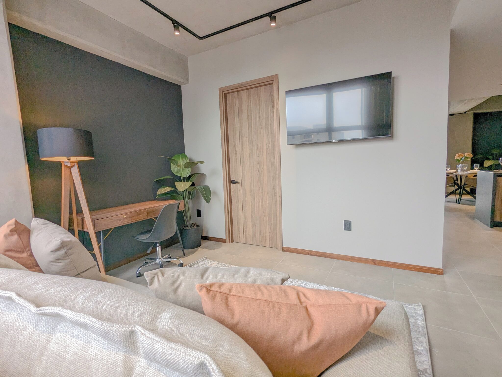 A modern bedroom with a bed, bedside table, lamp, desk, chair, potted plant, and a TV mounted on the wall.